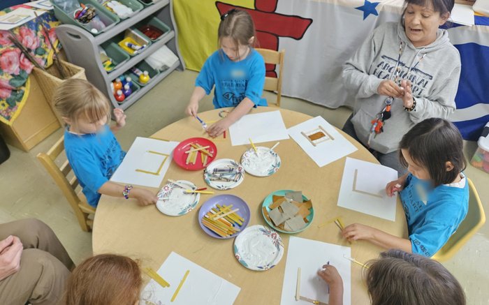 Children and a staff member around a craft table, gluing and painting craft sticks onto paper.