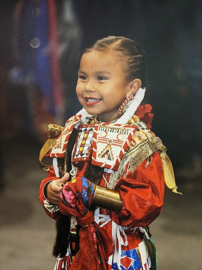A smiling young jingle dancer in a red beaded regalia dress at a powwow.