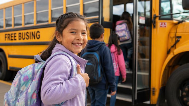 A smiling child with a backpack beside a yellow school bus marked Free Busing