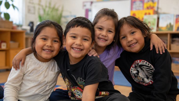 Four Head Start children with their arms around each other, smiling in a classroom