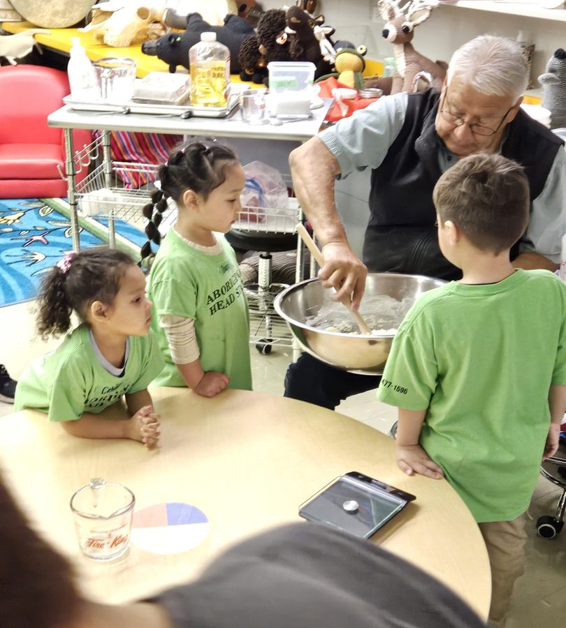 An Elder stirs a bowl of bannock dough while children watch from around the table.