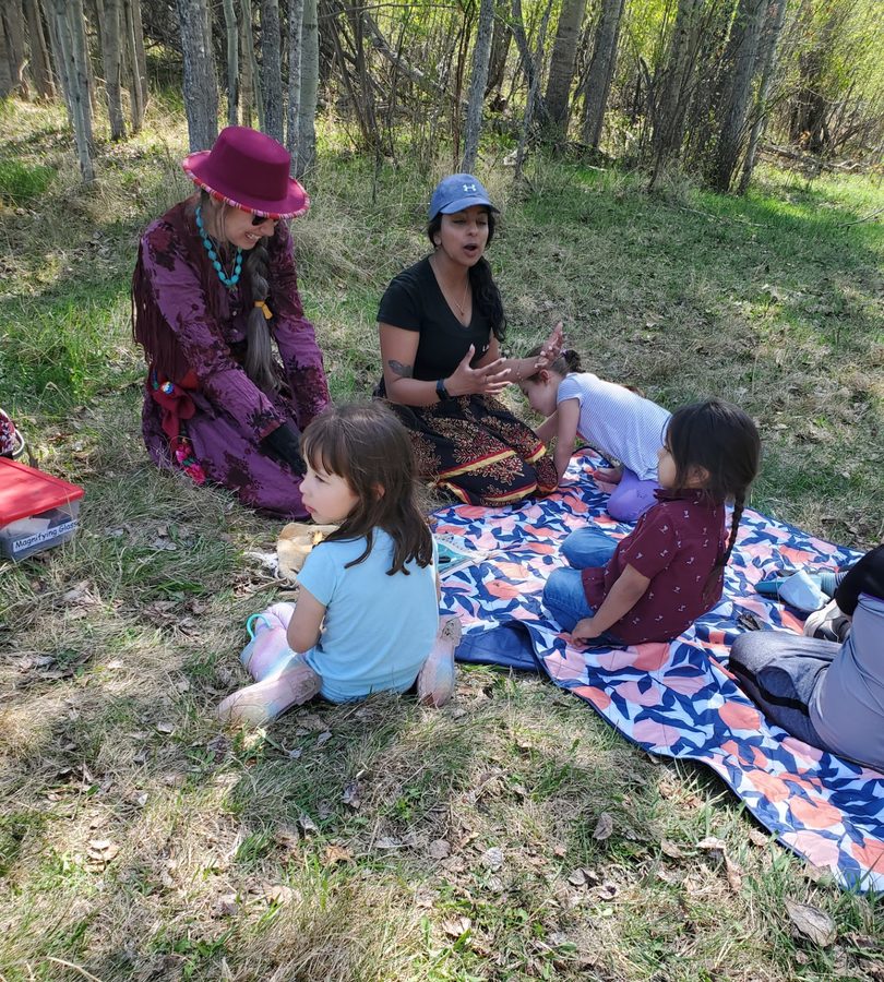 Children and two cultural mentors sitting together on a blanket in the trees.