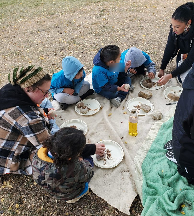 Children grinding dried meat with stones on plates, seated on a hide in the park.