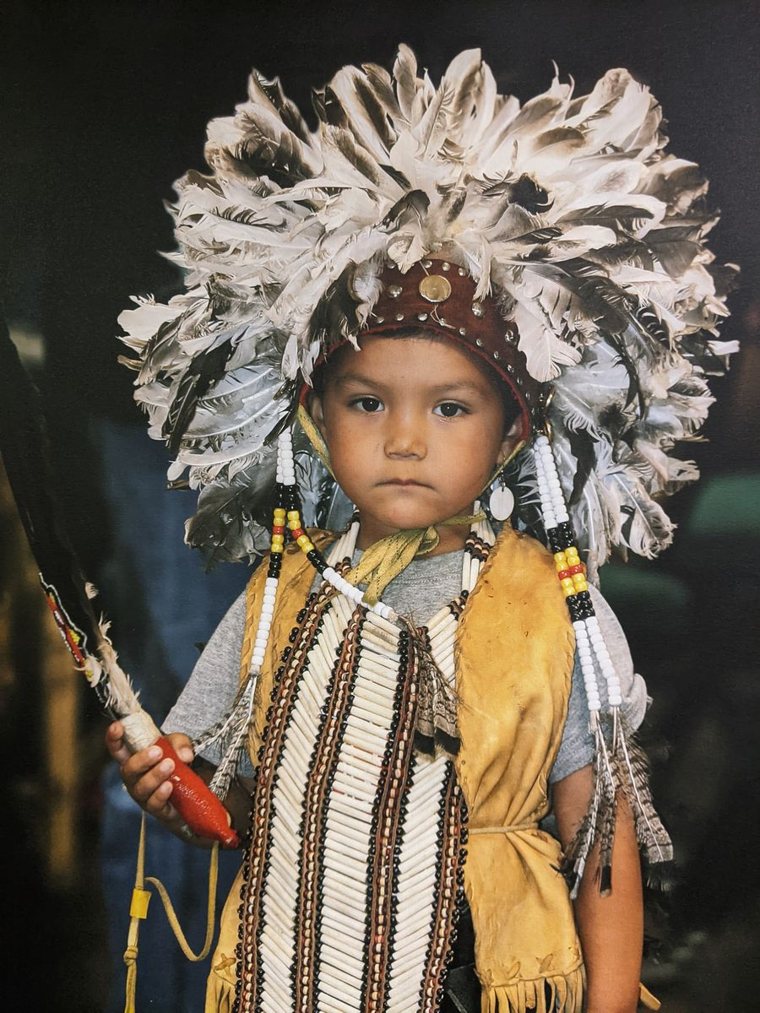 A young child in a feathered headdress and beaded regalia at a powwow.