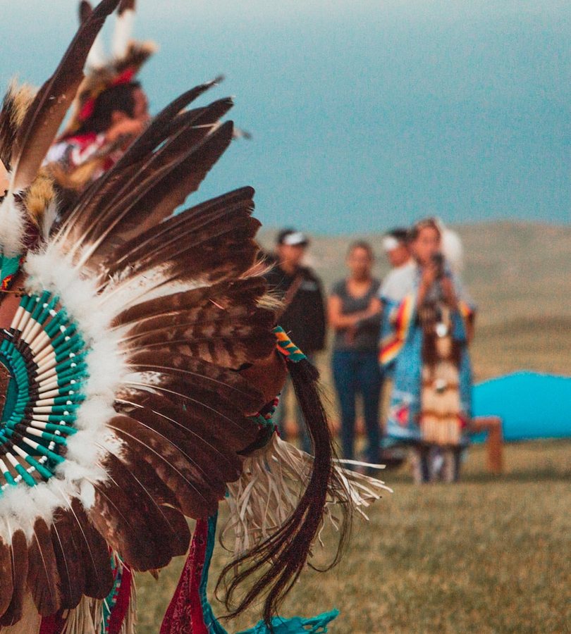 A dancer in a feathered bustle and beaded regalia at an outdoor powwow, with other dancers gathered on the grass behind