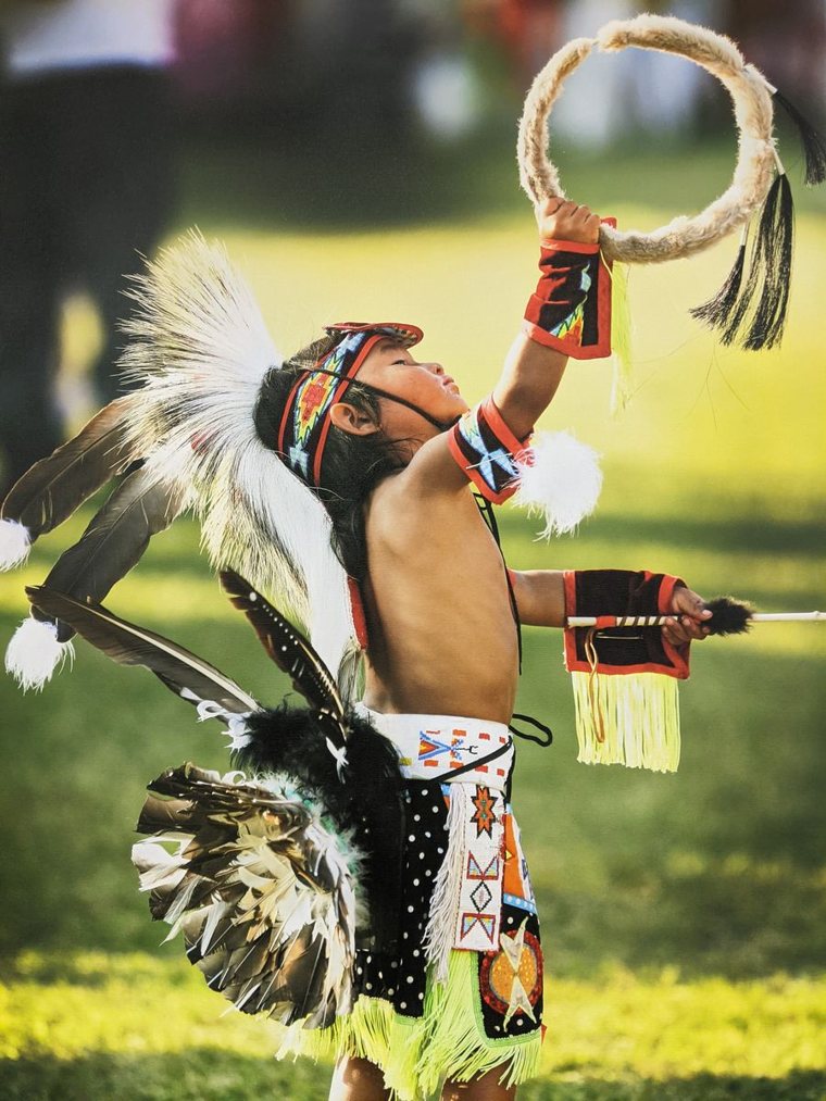 A young hoop dancer in beaded regalia and a feather bustle reaching up with a dance hoop at a powwow.