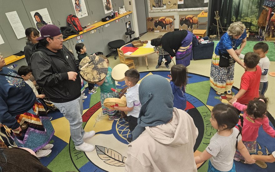 Children holding hand drums beside a drummer, standing on a medicine wheel rug