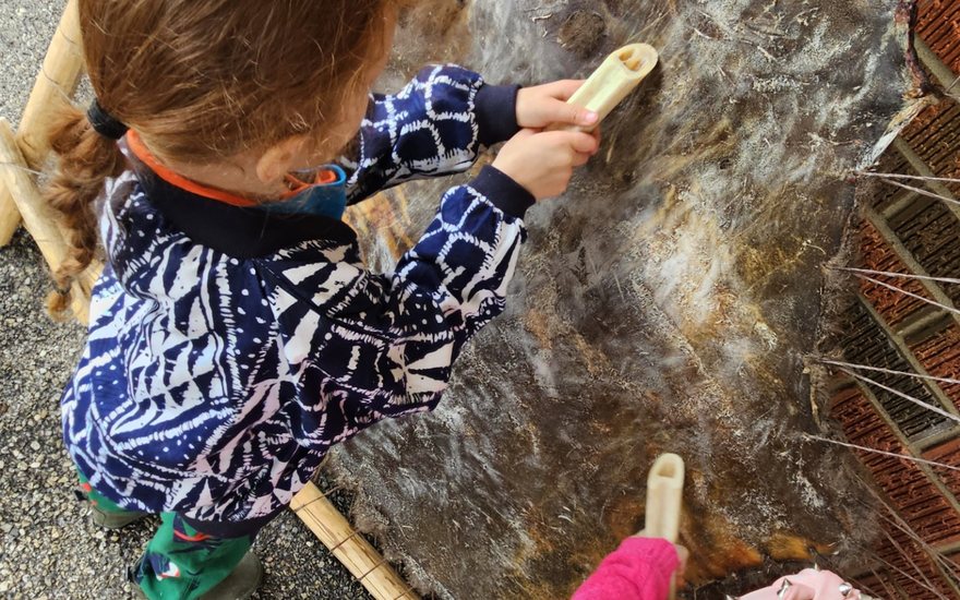 Two children using bone tools to scrape a hide stretched on a wooden frame